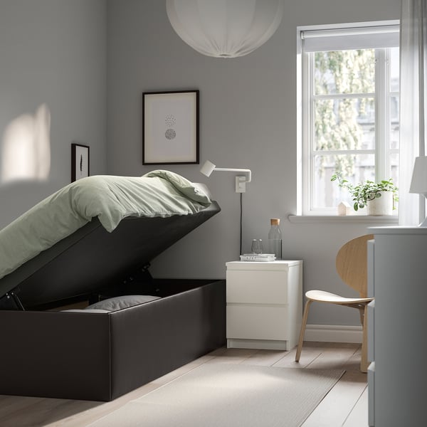 Modern bedroom with TJÅLLE storage bed open, showing internal space. Bed has light gray fabric and light wood floor. Nightstand with lamp beside bed.