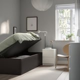 Modern bedroom with TJÅLLE storage bed open, showing internal space. Bed has light gray fabric and light wood floor. Nightstand with lamp beside bed.