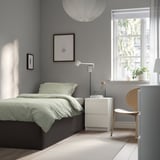 Modern bedroom with TJÅLLE bed, light green bedding, wooden chair, and white lamp. Natural light from large window, grey walls.