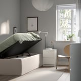 Minimalist bedroom with TJÅLLE storage bed, grey walls, white nightstand, light wood chair, and large window with sheer curtains.