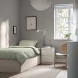 Sage green TJÅLLE bed with white nightstand, lamp, and flowers. Large window with sheer curtains, grey walls, wooden floors.