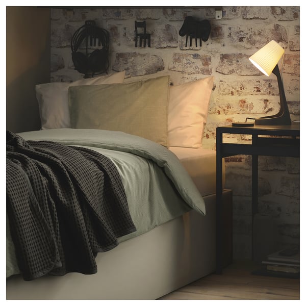Modern bed with light bedding, black blanket, and storage frame. Beside it, a lamp on a small table. Behind, a brick wall with decor.