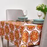Brightly colored tablecloth with sliced citrus patterns covers a table set with blue plates, wine glasses, and utensils, flanked by gray chairs.