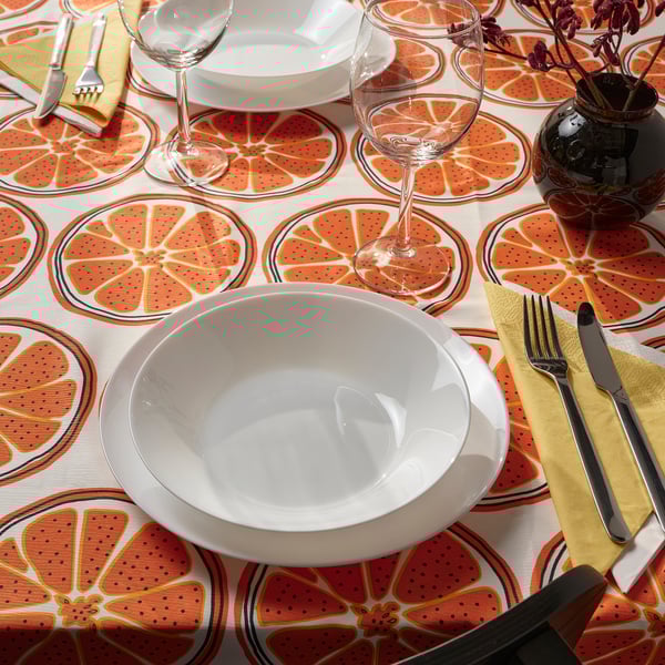 Bright orange citrus-patterned tablecloth with white bowl, two wine glasses, and yellow napkin, creating a cheerful dining setup.