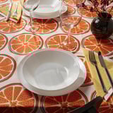 Bright orange citrus-patterned tablecloth with white bowl, two wine glasses, and yellow napkin, creating a cheerful dining setup.