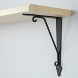 Wooden shelf mounted with black metal bracket, securely attached to wall, displaying simple & sturdy design.