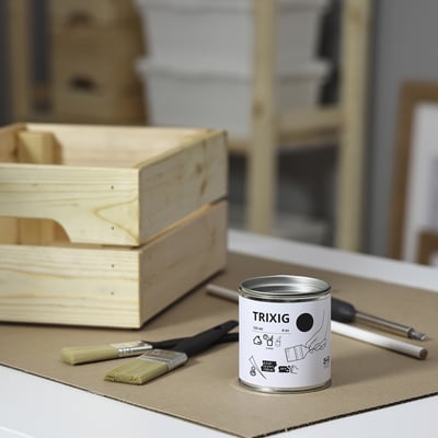 A can of TRIXIG black paint sits on cardboard, surrounded by brushes and a screwdriver, next to wooden crates.