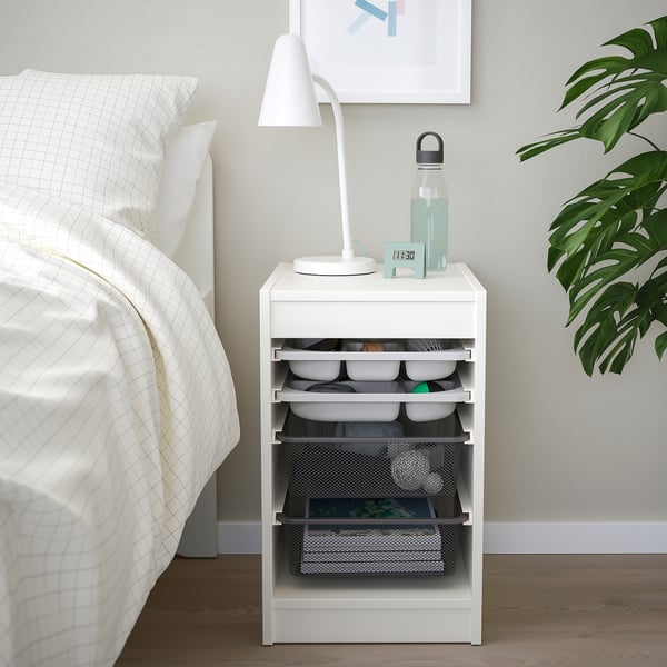 Modern white TROFAST storage unit beside bed, holding lamp, water bottle, and various containers.