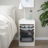 Modern white TROFAST storage unit beside bed, holding lamp, water bottle, and various containers.