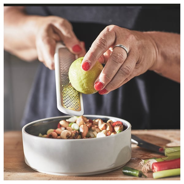 Person grates lime zest into bowl using small, white UPPFYLLD handheld grater. Bowl has mixed nuts and chopped veggies.