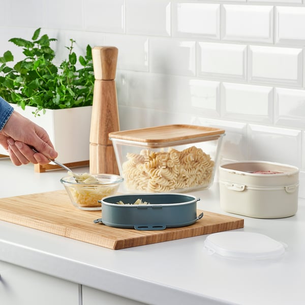 Blue stacking lunch box with snap-on lid, containing shredded cheese, placed on wooden board with other food containers and a hand taking some cheese.