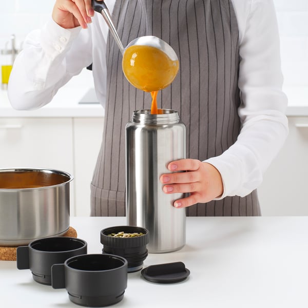 A person pours soup into a thermos on a counter with mugs and seed container.