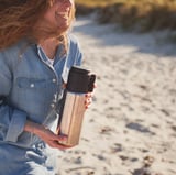 Woman holding a black and silver UTRUSTNING travel mug, smiling on a beach.