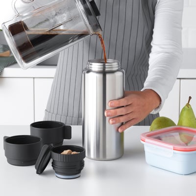Person pouring drink from pitcher into large insulated bottle with two mugs, ready for outdoor adventures.