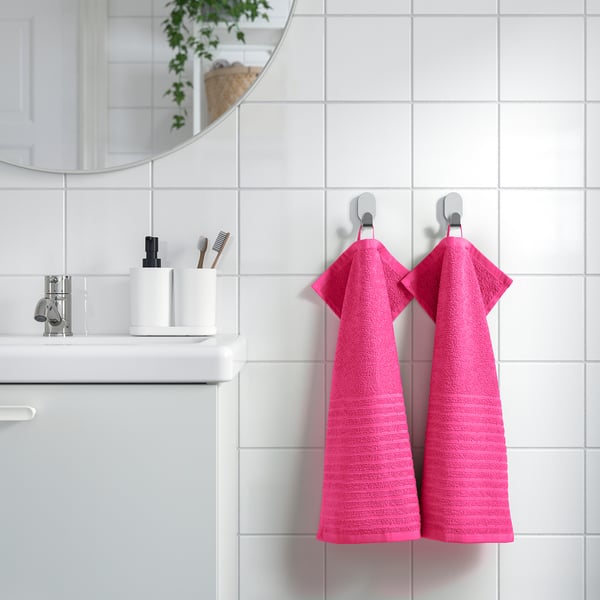 Bright pink VÅGSJÖN towel hangs beside bathroom sink, contrasting white tiles & accessories, small & compact.