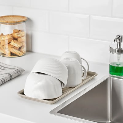 White dish drainer on kitchen counter next to sink, holding cups and bowls.