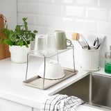 Kitchen counter with stainless steel sink, dish drainer holding cups and bowls, utensil holder, green plant, and cloth.