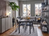 Modern dining room with extendable table, grey chairs, rug, plant, and shelves. Large windows with curtains.