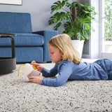 Child lies on carpet, playing with VAPPEBY speaker, its lamp on, blue sofa and plant nearby.