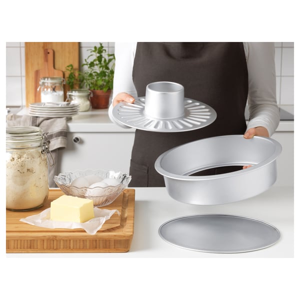 Person holding baking tins in kitchen. Various baking tools and ingredients visible. Tins have interchangeable bases for different cake types.