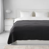 Bedroom with white bed, dark gray VÅRELD bedspread, white pillows, nightstand, lamp, clock, books, large rug.