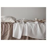 A white VÅRELD cotton bedspread on a single bed, adding warmth and decoration. Two pillows are at the head, with a mug on a tray beside them.