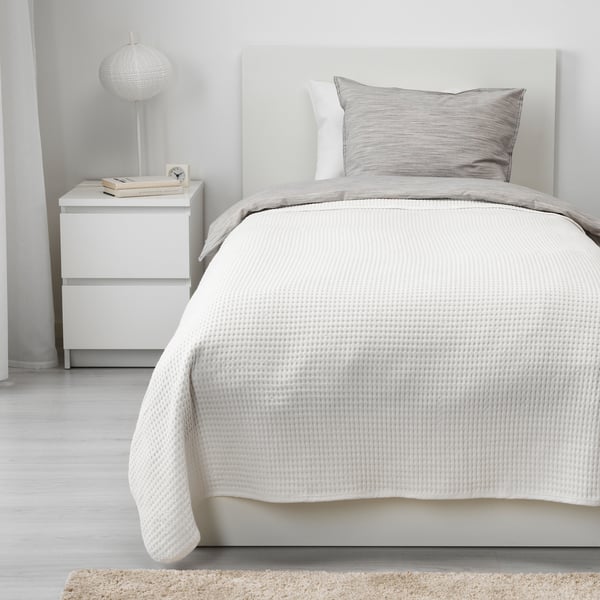 Cozy bedroom with white VÅRELD bed, soft cotton blanket. Nightstand with lamp & books, light walls & floor.