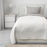 Cozy bedroom with white VÅRELD bed, soft cotton blanket. Nightstand with lamp & books, light walls & floor.
