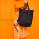 Woman holding black VÄRLDENS backpack, made of recycled polyester, with orange clothing against orange background.
