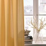 Yellow curtains hang by a window, partially covering it. Beside them, a vase with dry grasses sits on a stack of books.