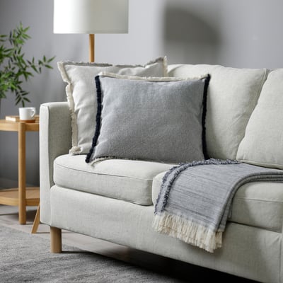 Modern living room with grey sofa, blue/white cushions, throw, lamp, side table, mug, plant.
