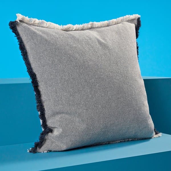 Grey VÄXELBRUK cushion with dark blue fringes, textured, against bright blue background.