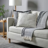VÄXELBRUK light grey cushion with fringes on sofa, smaller than nearby mug and lamp.