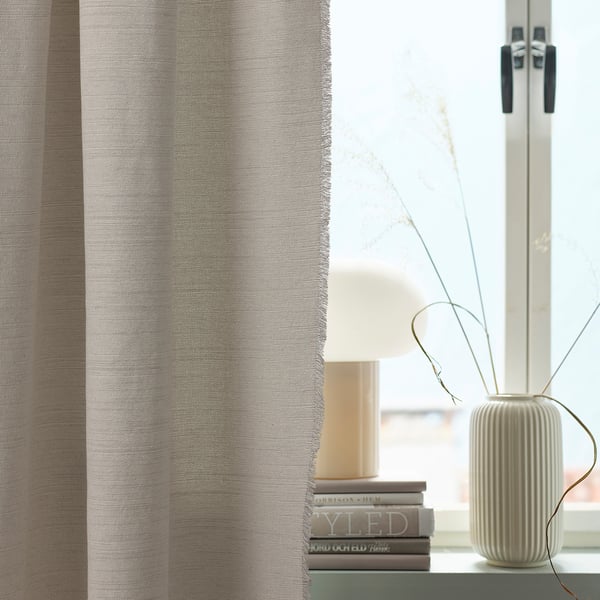Shared living room scene with grey VÄXELBRUK curtain, small lamp on books, tall white vase with branches.