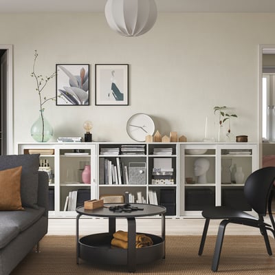 Minimalist living room with gray storage, glass doors, books, vases, decor. Wall hangings, pendant light, gray sofa, black chair, coffee table, rug.