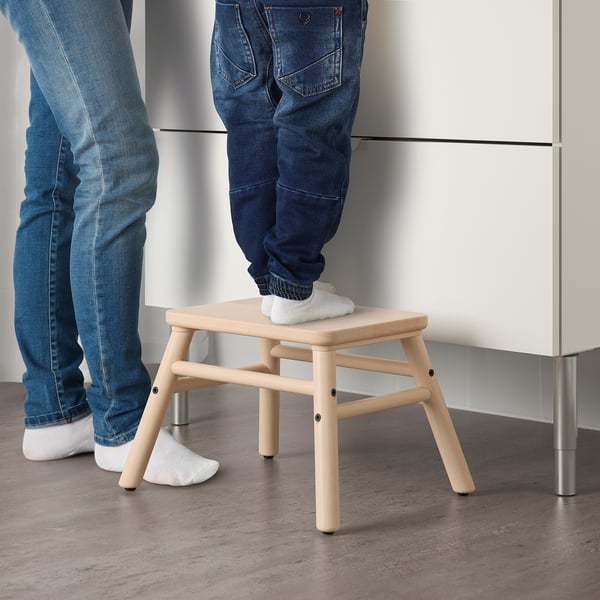 Two people use a wooden stool, one standing on it, likely to reach higher areas, showcasing its function as a step stool.