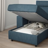 Blue storage sofa bed, open compartment showing folded sheets, white wall behind.