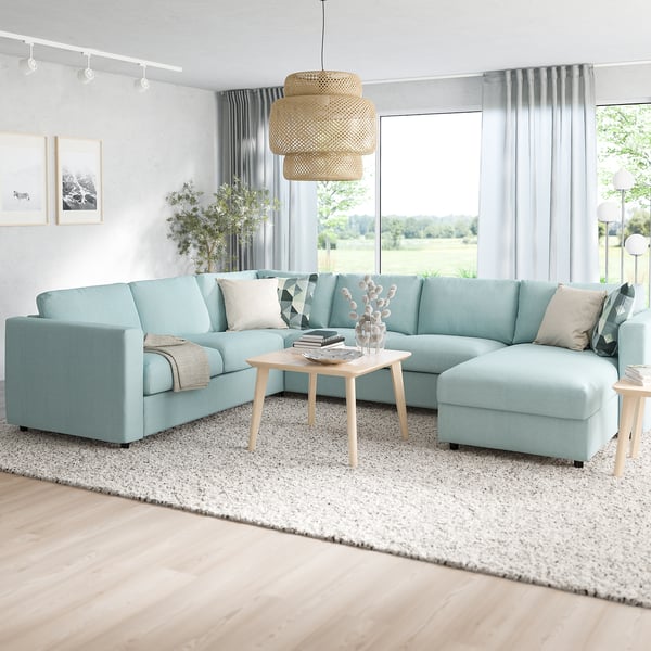 Modern living room with light blue l-shaped VIMLE sofa, wooden coffee table, white curtains, and large windows. Overhead lights and a textured rug.
