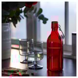 Red glass bottle with stopper next to clear glass on a table.