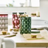 Three festive tin containers on wooden counter with treats and holiday decorations.
