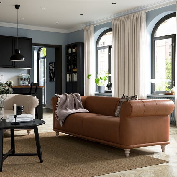 Modern living room with brown leather VISKAFORS sofa, wooden side table, black pendant lamp, arched windows, and black bookshelf.