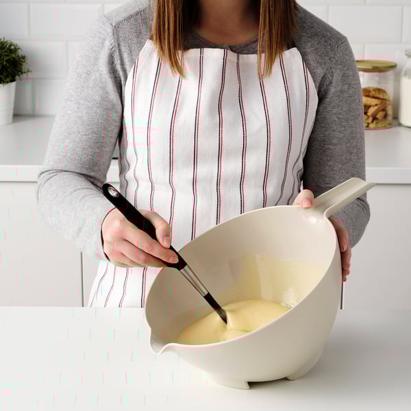 Person holding a mixing bowl with a spout for easy pouring, wearing an apron in a kitchen.
