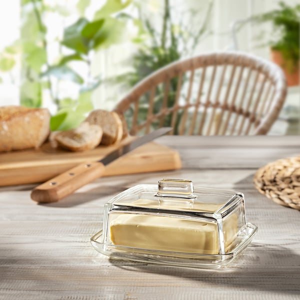 Glass butter dish with lid, yellow butter, cutting boards, knife, bread, wicker chair.