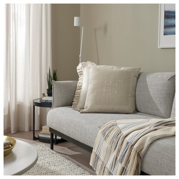 Acogedora sala de estar con cojín ÅKERNEJLIKA beige sobre sofá gris claro, con flores bordadas a mano.
