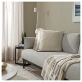 Acogedora sala de estar con cojín ÅKERNEJLIKA beige sobre sofá gris claro, con flores bordadas a mano.