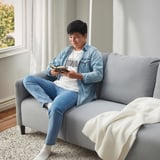 Un joven está sentado en un sofá gris de ANGERSBY, leyendo un libro. Suave, cómodo y práctico con bolsillos laterales.