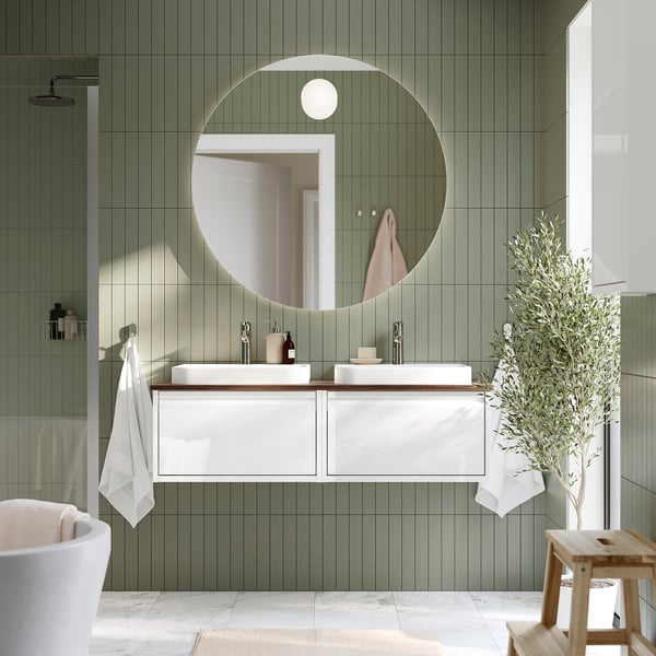 Moderno baño con azulejos verdes con gran espejo redondo, tocador doble de mármol blanco y taburete de madera.