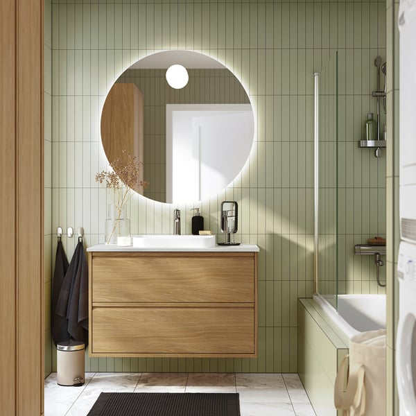 Baño moderno con tocador de roble ÄNGSJÖN, azulejos verdes, ducha de vidrio y toalla beige.