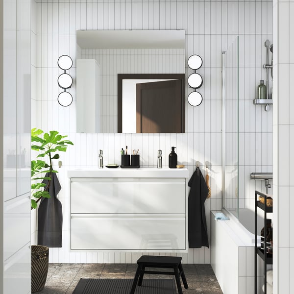 Moderno baño blanco con gabinete de lavabo ÄNGSJÖN, tazón doble, detalles en negro y lámparas redondas.