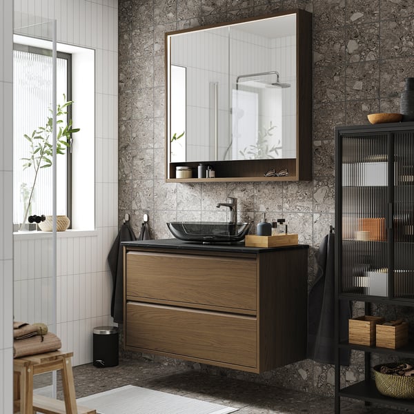 Baño moderno con muebles ÄNGSJÖN de roble marrón. Tocador montado en la pared, espejo grande, almacenamiento alto y taburete de madera.